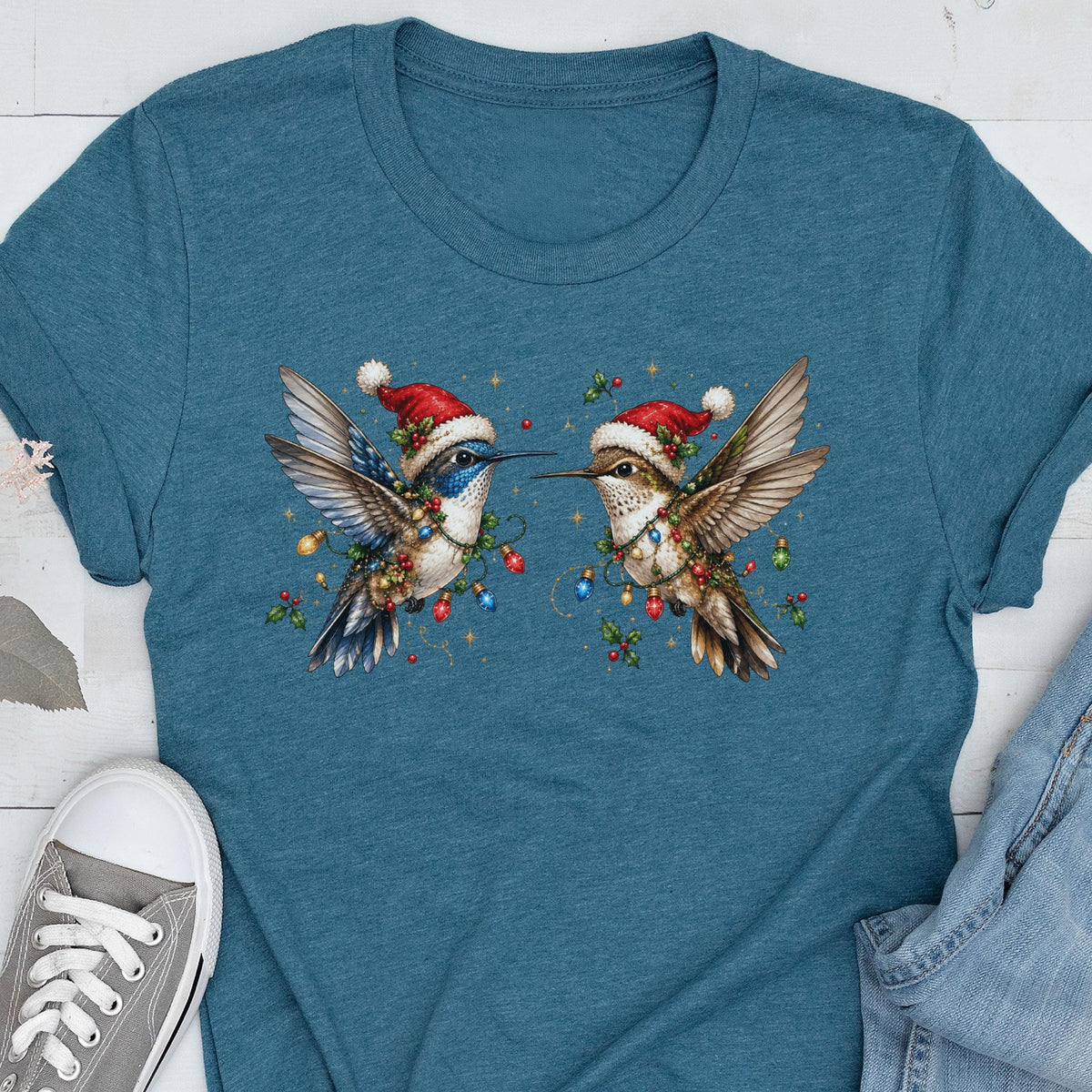 May include: A teal blue short-sleeve t-shirt featuring a graphic of two hummingbirds facing each other. Each bird wears a red and white Santa hat and is wrapped in colorful Christmas light bulbs and green holly leaves with red berries.