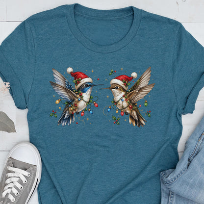 May include: A teal blue short-sleeve t-shirt featuring a graphic of two hummingbirds facing each other. Each bird wears a red and white Santa hat and is wrapped in colorful Christmas light bulbs and green holly leaves with red berries.