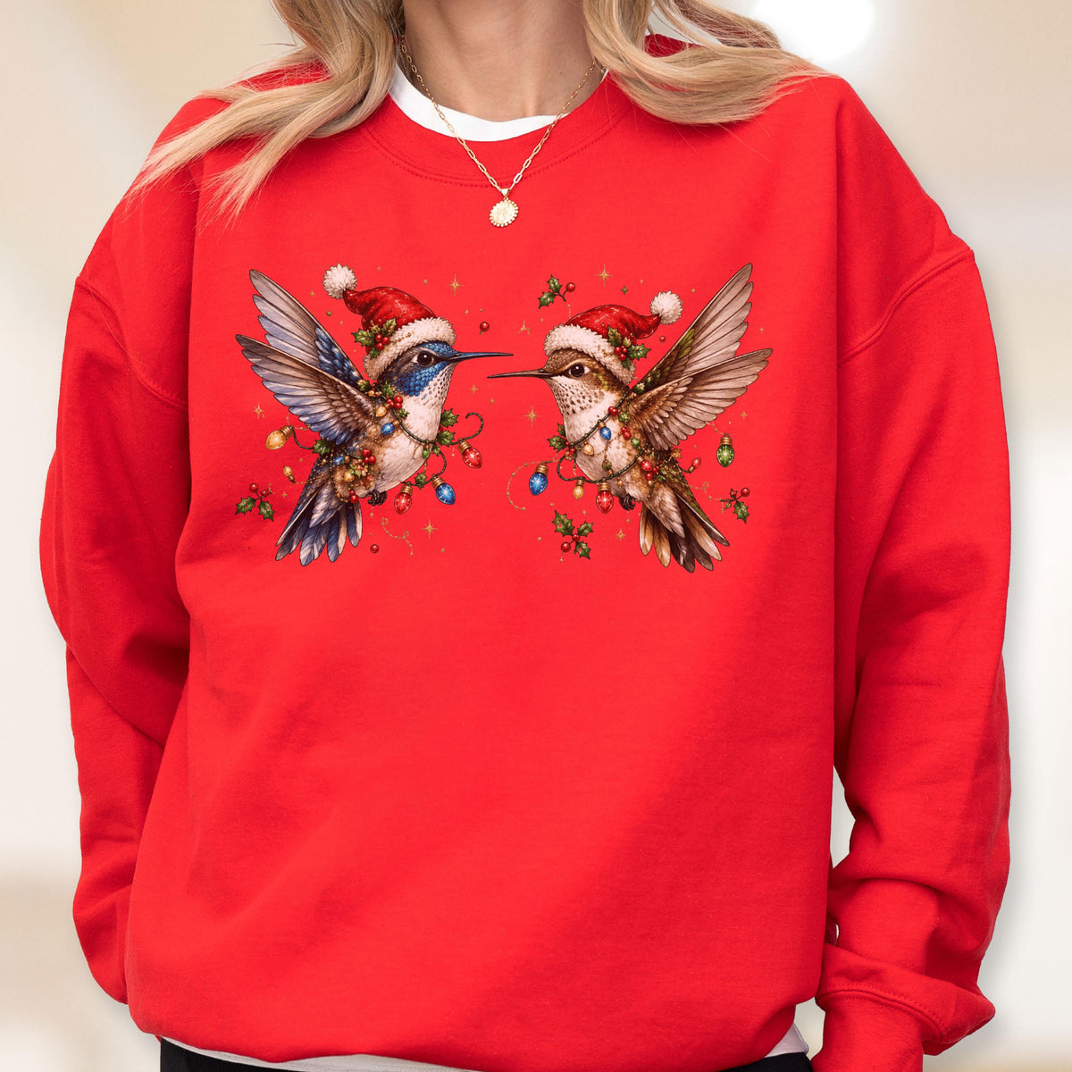 May include: A bright red crewneck sweatshirt featuring a graphic of two hummingbirds facing each other. Each bird wears a small red and white Santa hat and is surrounded by colorful Christmas light bulbs, green holly leaves, and small red berries.
