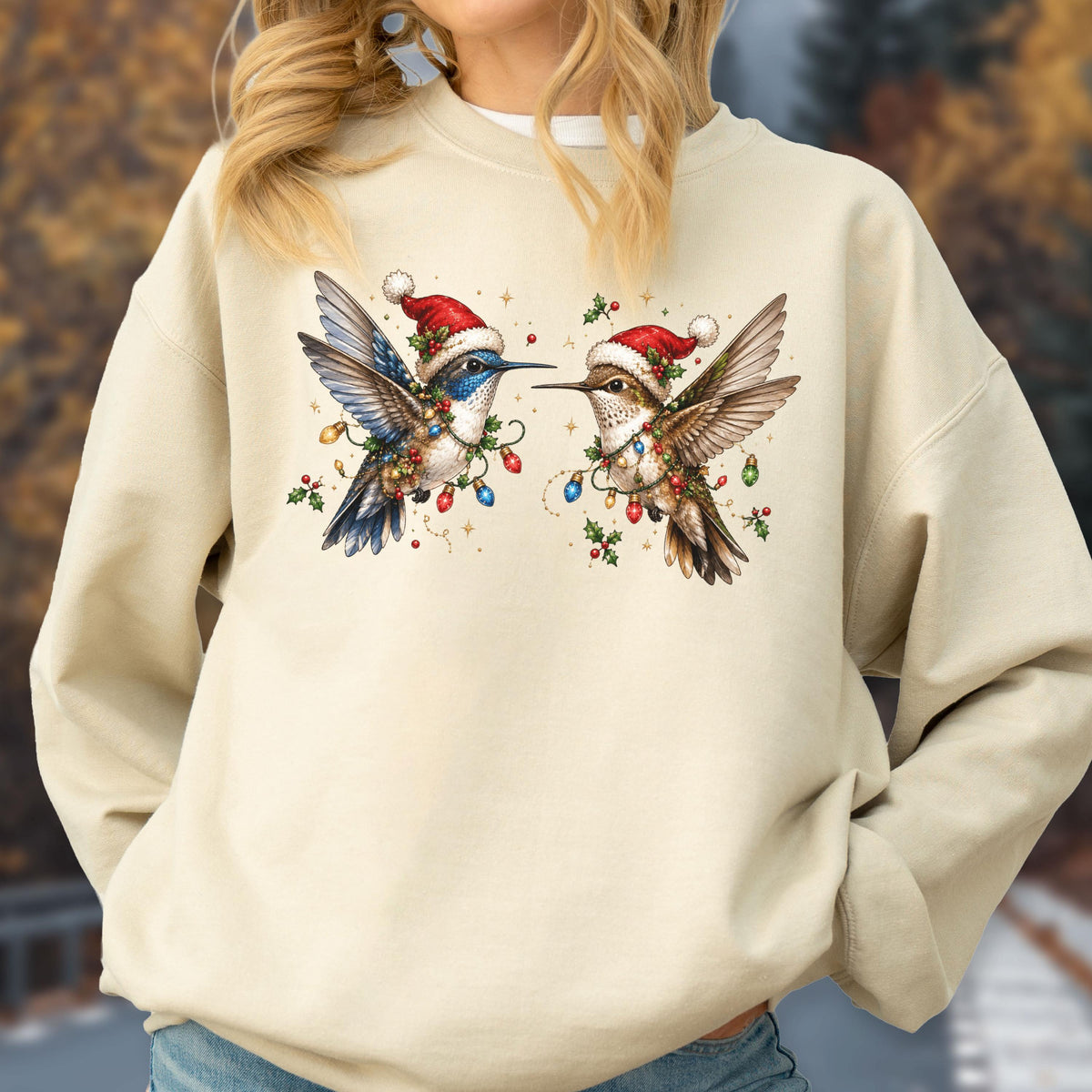 May include: A beige crewneck sweatshirt featuring a graphic print of two hummingbirds wearing red and white Santa hats. The birds are surrounded by colorful Christmas light bulbs, green holly leaves, and small red berries, creating a festive scene.