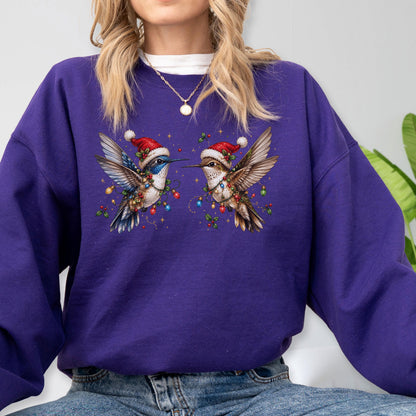 May include: A deep purple long-sleeve sweatshirt featuring a graphic of two hummingbirds facing each other. Each bird wears a red Santa hat and is wrapped in colorful Christmas lights with small green holly leaves and berries scattered around.