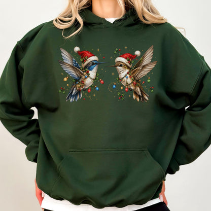 May include: A dark forest green hooded sweatshirt featuring a graphic print of two hummingbirds wearing red Santa hats. The birds are adorned with colorful Christmas light strings and holly berries, set against a dark green fabric background.