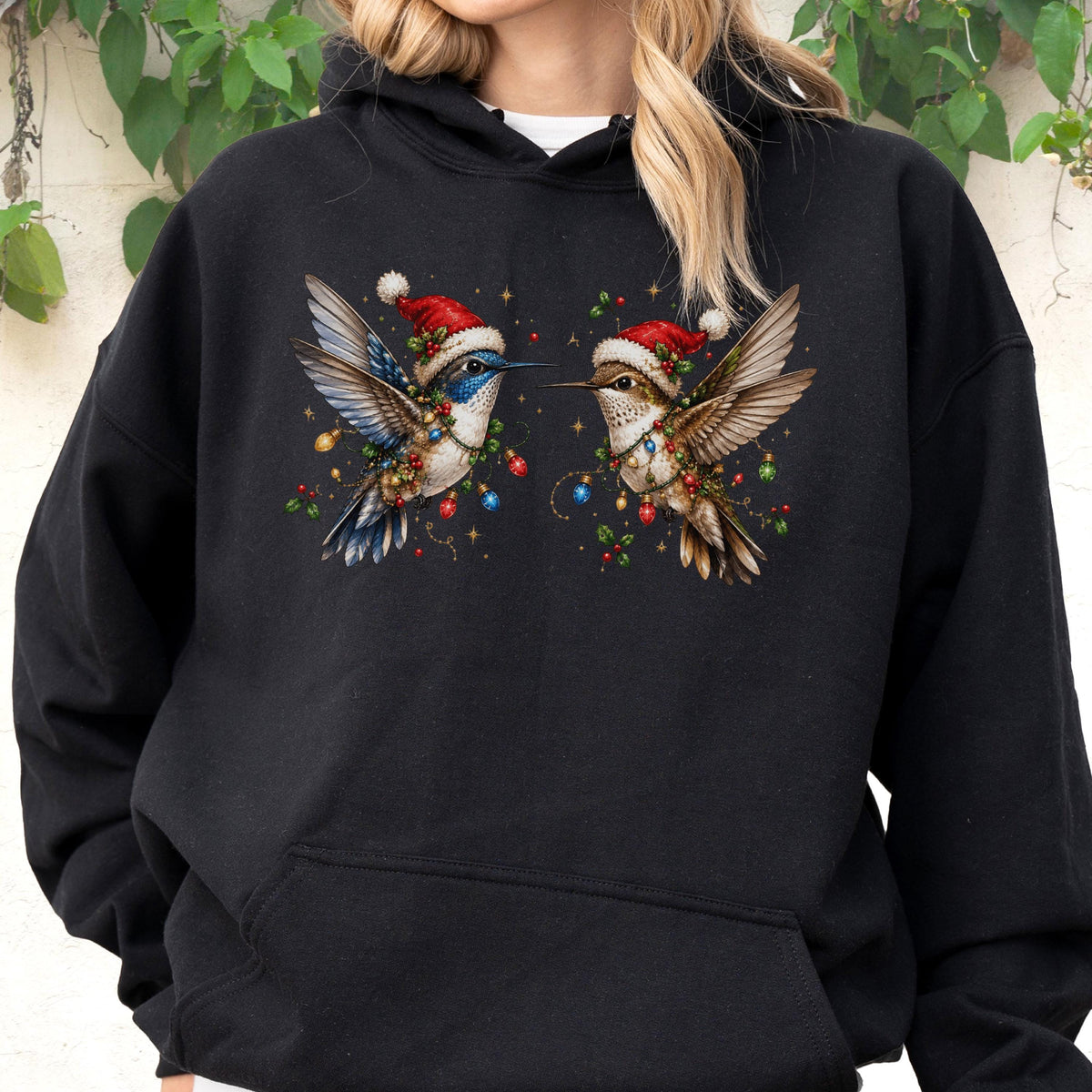 May include: A black hooded sweatshirt featuring a graphic print of two hummingbirds facing each other. Each bird wears a red and white Santa hat and is wrapped in colorful Christmas lights with small red berries and green leaves around them.