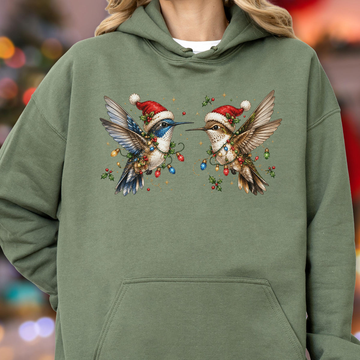May include: An olive green hooded sweatshirt featuring a graphic print of two hummingbirds wearing red and white Santa hats. The birds are surrounded by colorful Christmas light bulbs and green holly leaves with red berries on the chest area.