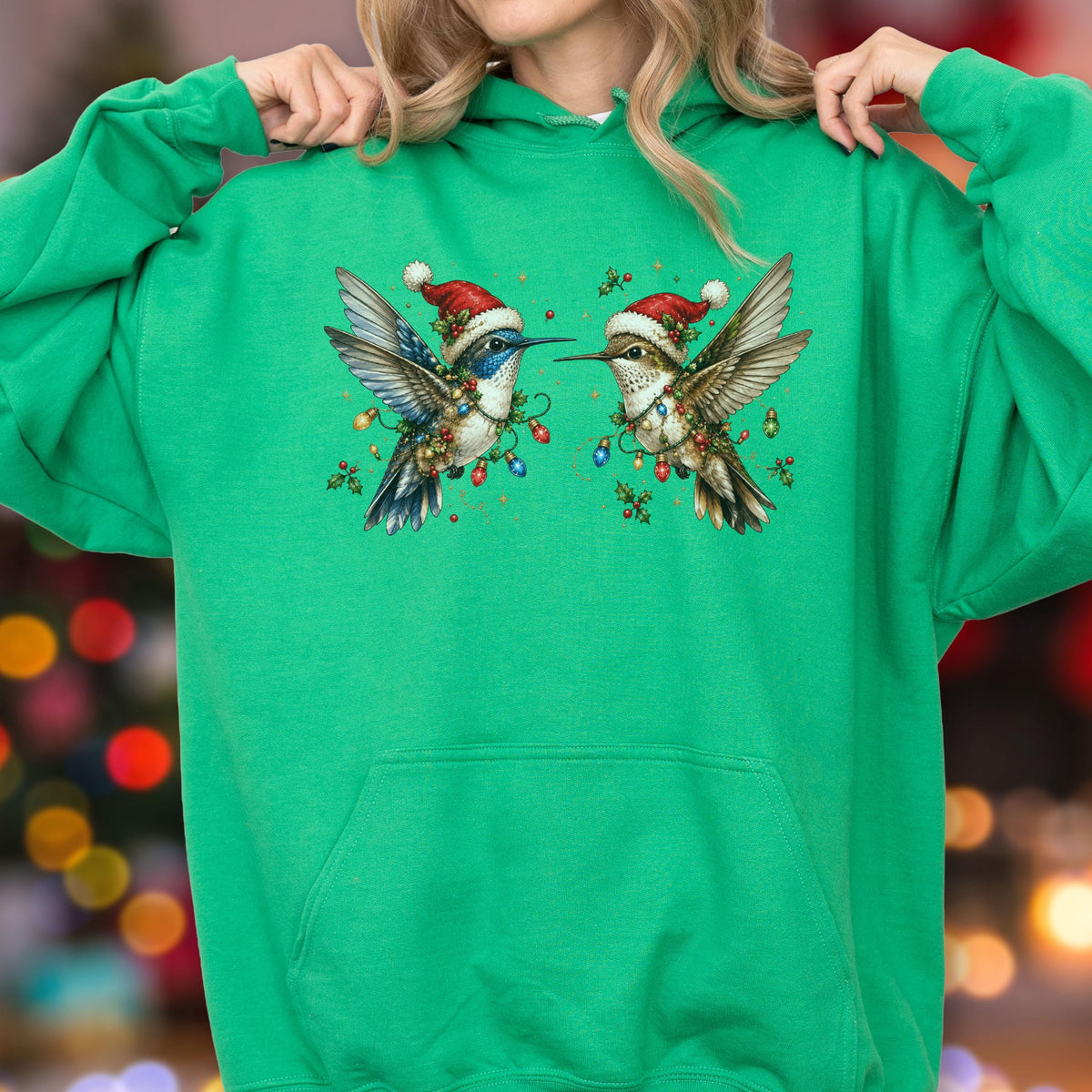 May include: A bright green hooded sweatshirt featuring a graphic of two hummingbirds wearing red and white Santa hats. The birds are decorated with colorful Christmas light strings and green holly leaves, centered on the front of the garment.