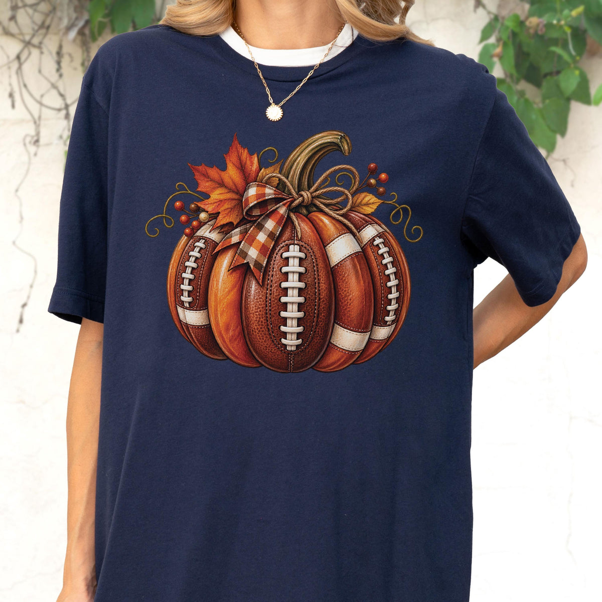 May include: A navy blue short-sleeve t-shirt featuring a graphic of a pumpkin designed to look like a brown leather football with white laces. The pumpkin is decorated with a plaid ribbon bow, orange maple leaves, and small red berries.
