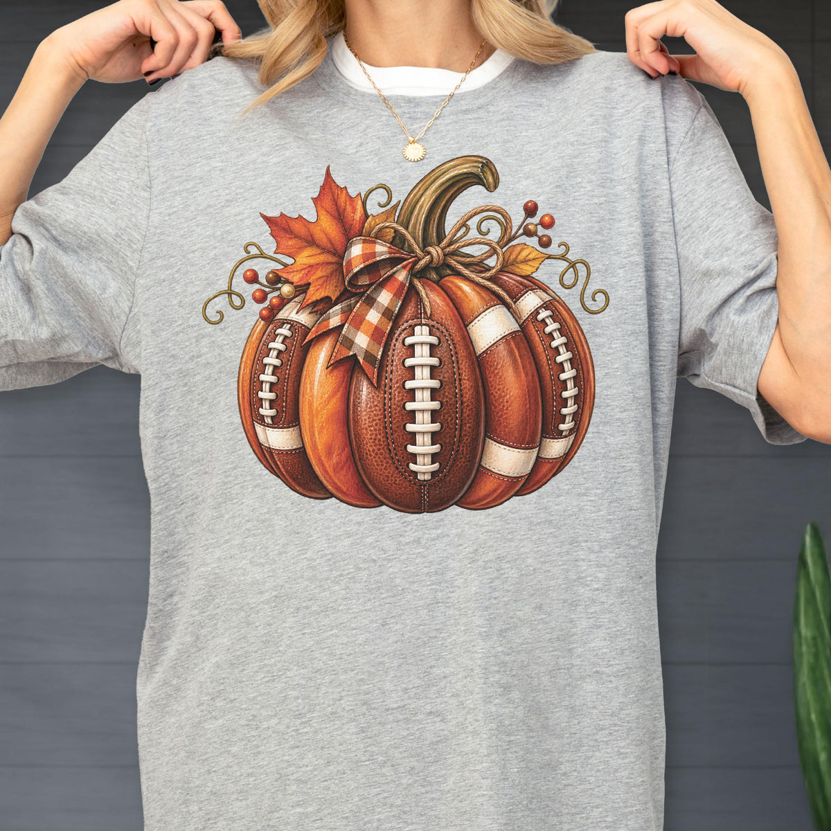 May include: A heather gray t-shirt featuring a graphic of a pumpkin designed with the texture and white laces of a football. The pumpkin is decorated with a plaid ribbon bow, orange maple leaves, and small brown berries on a vine-like stem.