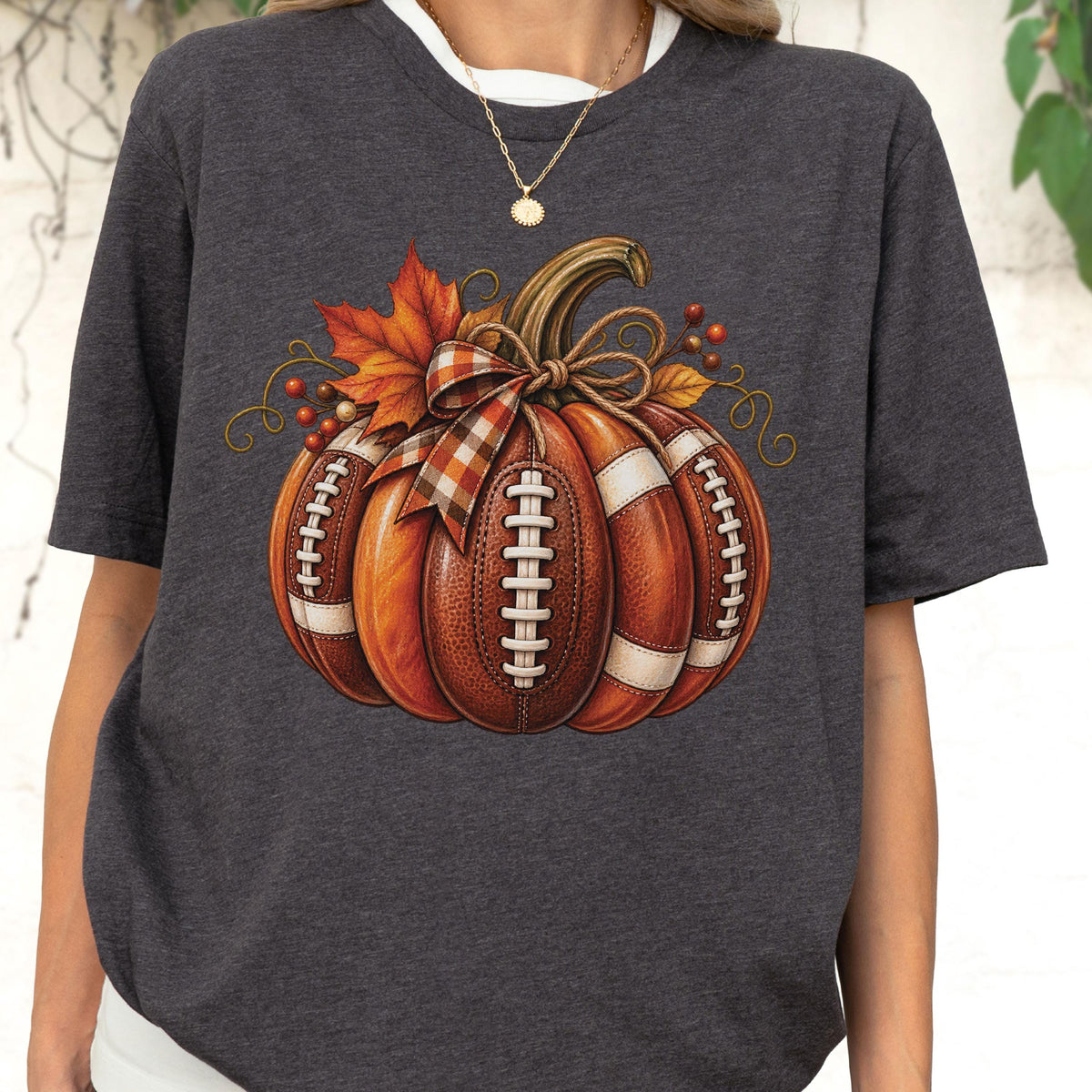 May include: A dark charcoal gray short-sleeve t-shirt featuring a central graphic of a pumpkin designed with the texture and white laces of an American football. The pumpkin is adorned with a plaid ribbon bow, orange maple leaves, and berries.