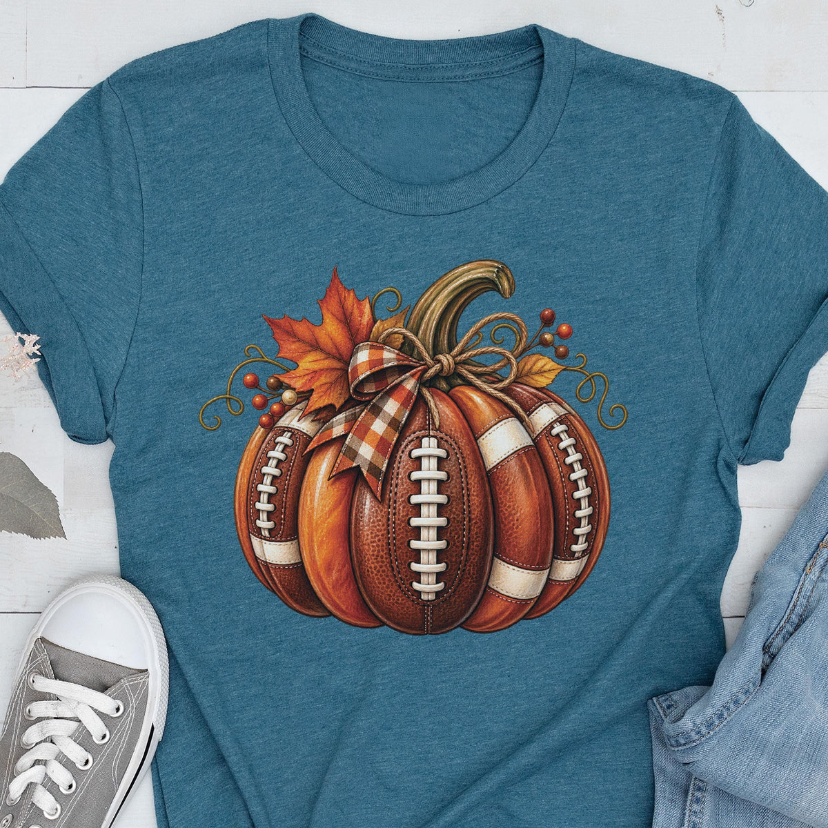May include: A heathered teal short-sleeve t-shirt featuring a central graphic of a pumpkin designed with the texture and white laces of an American football. The pumpkin is decorated with a plaid ribbon bow, orange maple leaves, and small berries.
