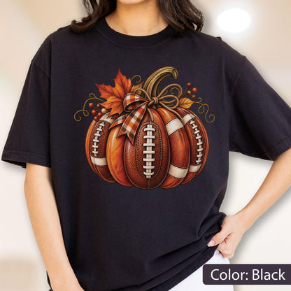 May include: A black short-sleeved t-shirt featuring a graphic of a pumpkin designed to look like a brown leather football with white laces. The pumpkin is decorated with a plaid bow, orange maple leaves, and small red berries on top.