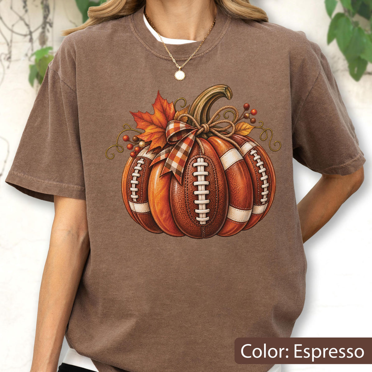 May include: An espresso brown t-shirt featuring a graphic of a pumpkin designed with the texture and white laces of a football. The pumpkin is decorated with a plaid ribbon bow, orange autumn leaves, and small red berries on its stem.
