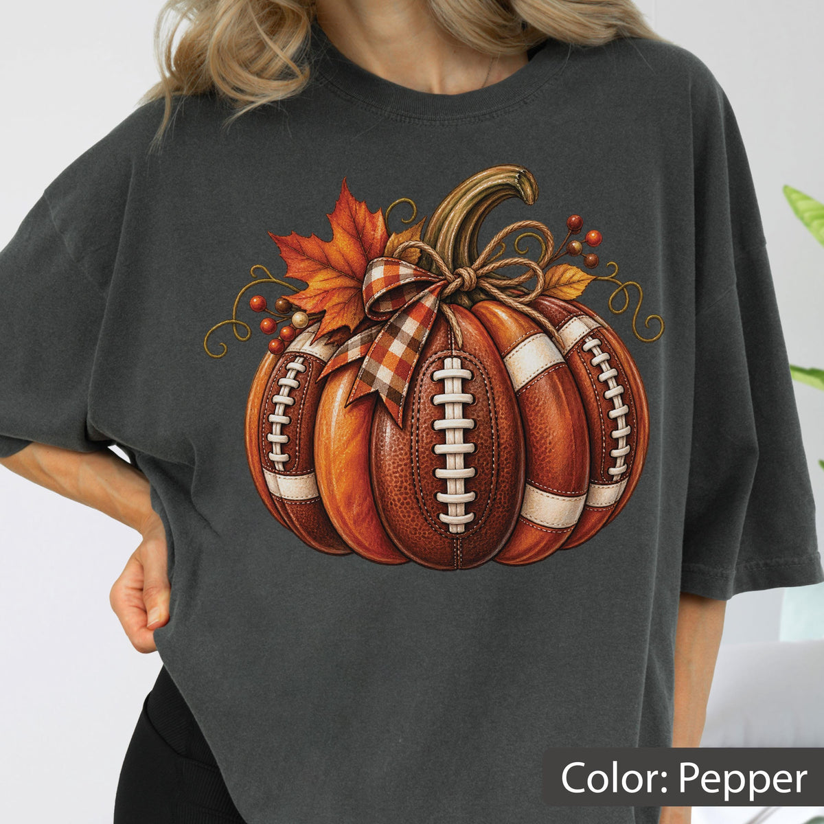 May include: A charcoal gray t-shirt featuring a graphic of a pumpkin designed with the texture and white laces of a football. The pumpkin is decorated with a plaid ribbon bow, orange autumn leaves, and small red berries on the top stem.