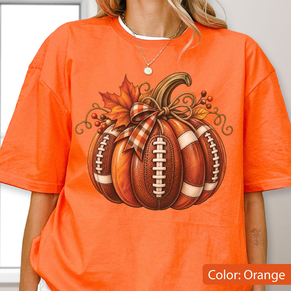 May include: An orange short-sleeve t-shirt featuring a graphic of a pumpkin designed with the texture and white laces of an American football. The pumpkin is decorated with a plaid ribbon, autumn leaves, and small berries on a vine.
