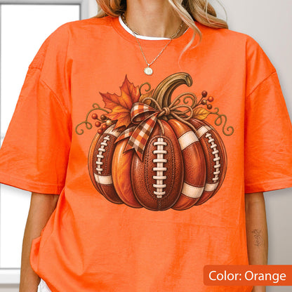 May include: An orange short-sleeve t-shirt featuring a graphic of a pumpkin designed with the texture and white laces of an American football. The pumpkin is decorated with a plaid ribbon, autumn leaves, and small berries on a vine.