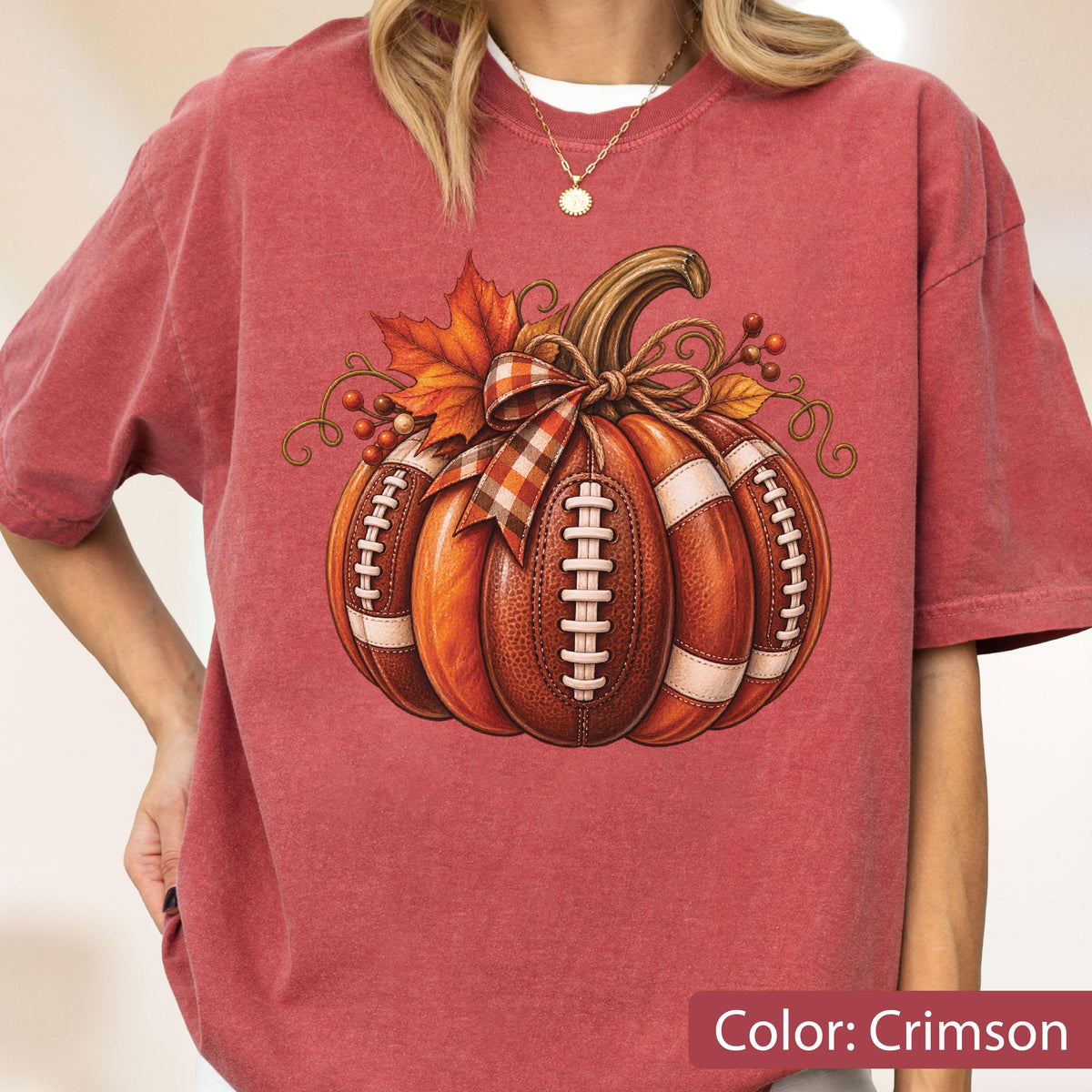 May include: A crimson short-sleeve t-shirt featuring a graphic of a pumpkin designed to look like a brown leather football with white laces. The pumpkin is decorated with a plaid ribbon bow, orange autumn leaves, and small red berries.