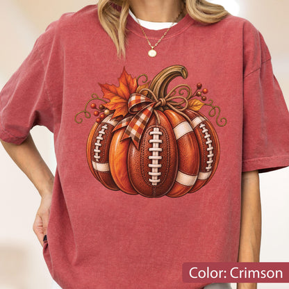 May include: A crimson short-sleeve t-shirt featuring a graphic of a pumpkin designed to look like a brown leather football with white laces. The pumpkin is decorated with a plaid ribbon bow, orange autumn leaves, and small red berries.