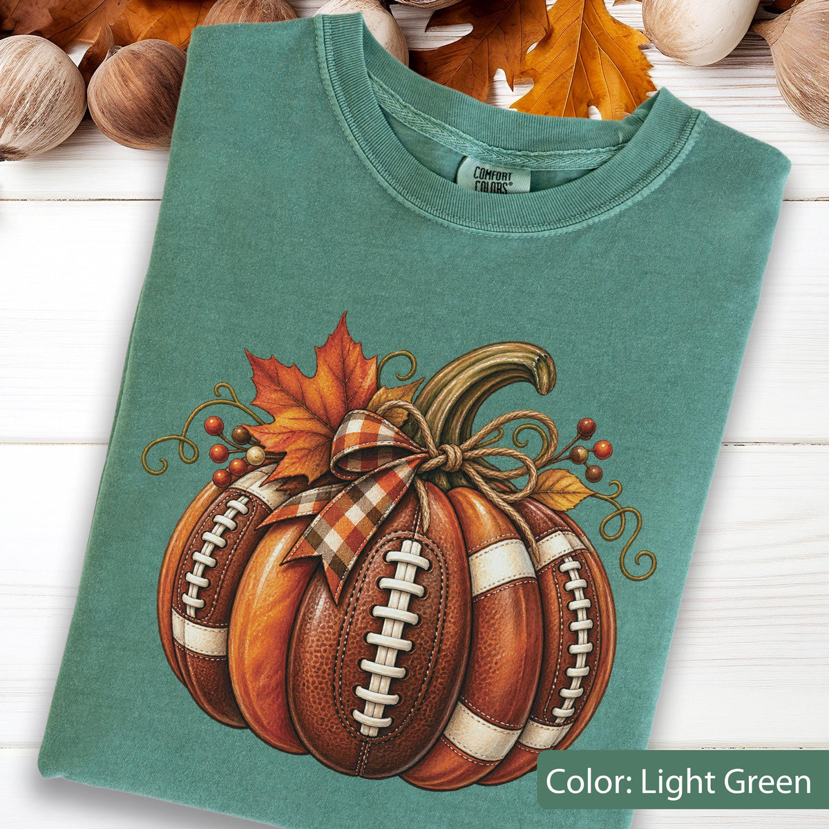May include: A light green crew neck t-shirt featuring a graphic of a pumpkin designed with brown football leather textures, white laces, and a plaid ribbon bow. The design includes an orange maple leaf and small red berries with curly vines.