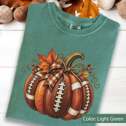 May include: A light green crew neck t-shirt featuring a graphic of a pumpkin designed with brown football leather textures, white laces, and a plaid ribbon bow. The design includes an orange maple leaf and small red berries with curly vines.