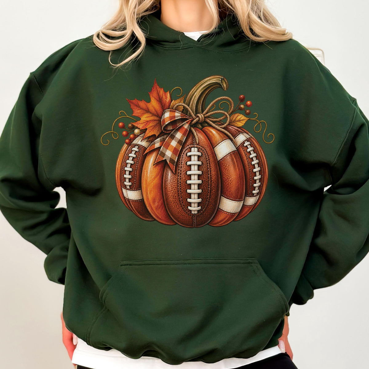 May include: A dark forest green hooded sweatshirt featuring a central graphic of a pumpkin constructed from brown footballs with white laces. The pumpkin is decorated with a plaid ribbon bow, orange maple leaves, and small red berries.