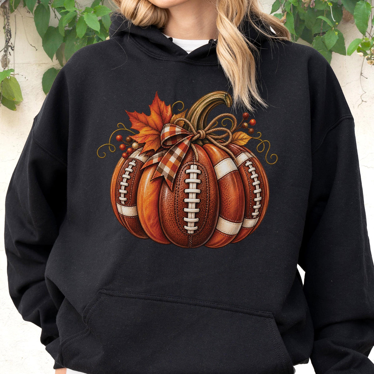 May include: A black hooded sweatshirt featuring a graphic print of a pumpkin designed with the texture and white laces of an American football. The pumpkin is decorated with a plaid ribbon bow, orange autumn leaves, and small red berries.