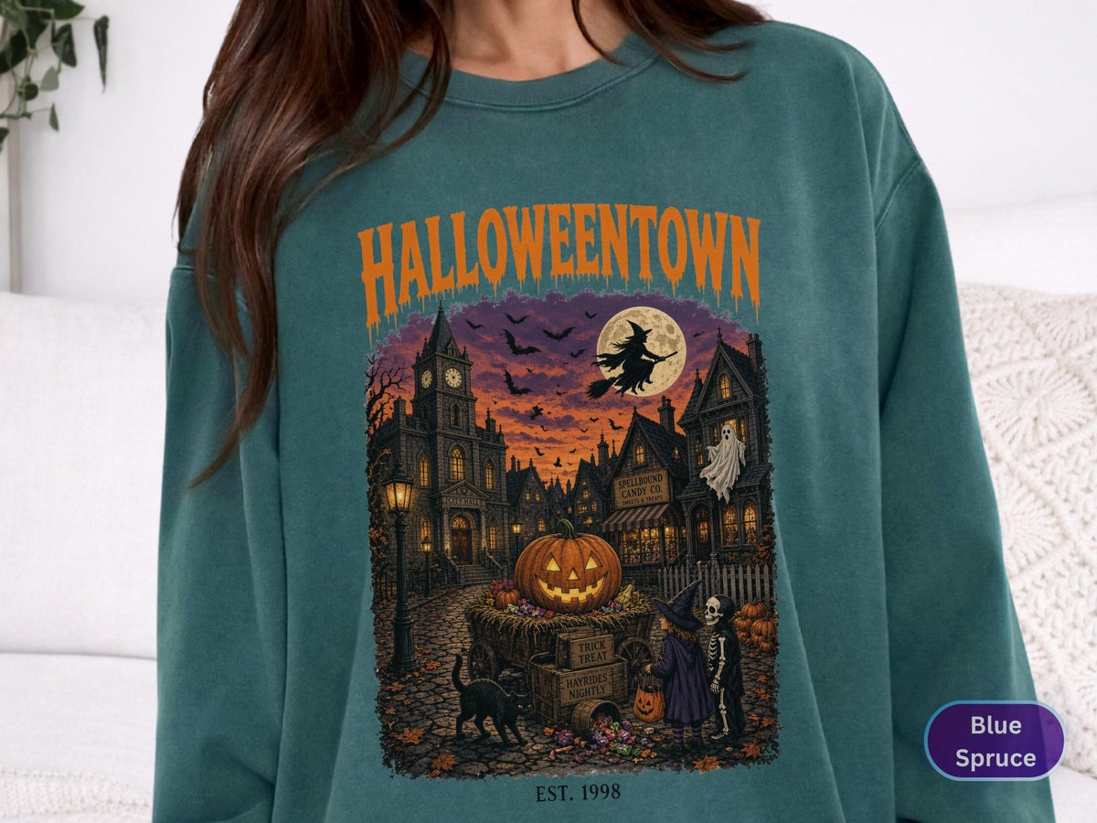 May include: A blue spruce crewneck sweatshirt featuring a graphic print with the orange text HALLOWEENTOWN and EST. 1998. The illustration depicts a spooky town scene at night with a large pumpkin, a witch on a broom, a ghost, and children.