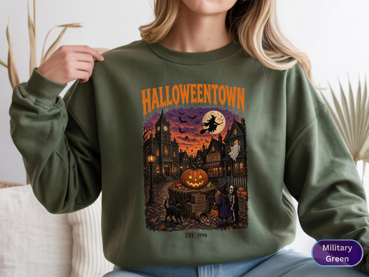 May include: A military green crewneck sweatshirt featuring a graphic print with the text HALLOWEENTOWN and EST. 1998. The illustration depicts a spooky town scene with a large jack-o'-lantern, a witch on a broomstick, a ghost, and a black cat.