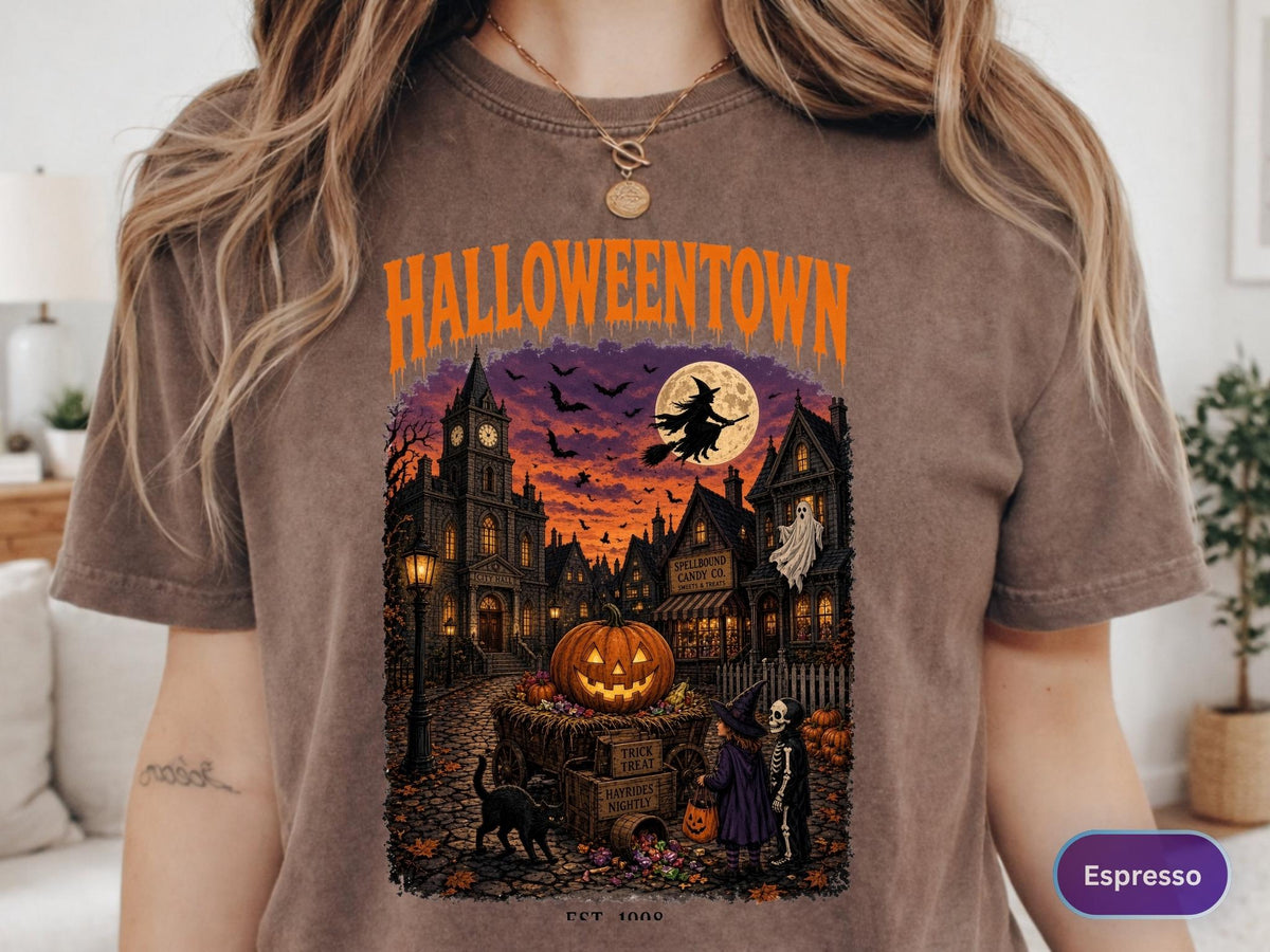 May include: A brown t-shirt featuring a graphic print with the text HALLOWEENTOWN in orange. The illustration depicts a spooky town scene at sunset with a large pumpkin, a flying witch, a ghost, a black cat, and two children in costumes.