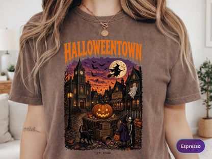 May include: A brown t-shirt featuring a graphic print with the text HALLOWEENTOWN in orange. The illustration depicts a spooky town scene at sunset with a large pumpkin, a flying witch, a ghost, a black cat, and two children in costumes.