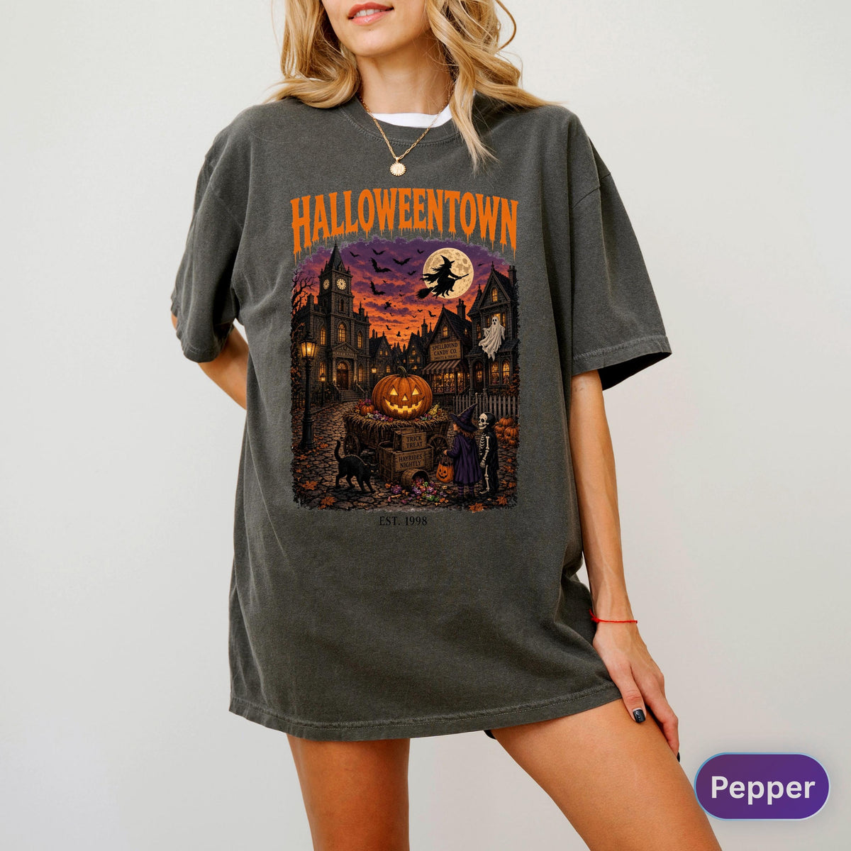 May include: A charcoal gray short-sleeve t-shirt featuring a graphic print with the text HALLOWEENTOWN and EST. 1998. The design depicts a spooky village scene at night with a large orange pumpkin, a flying witch, a ghost, and a black cat.