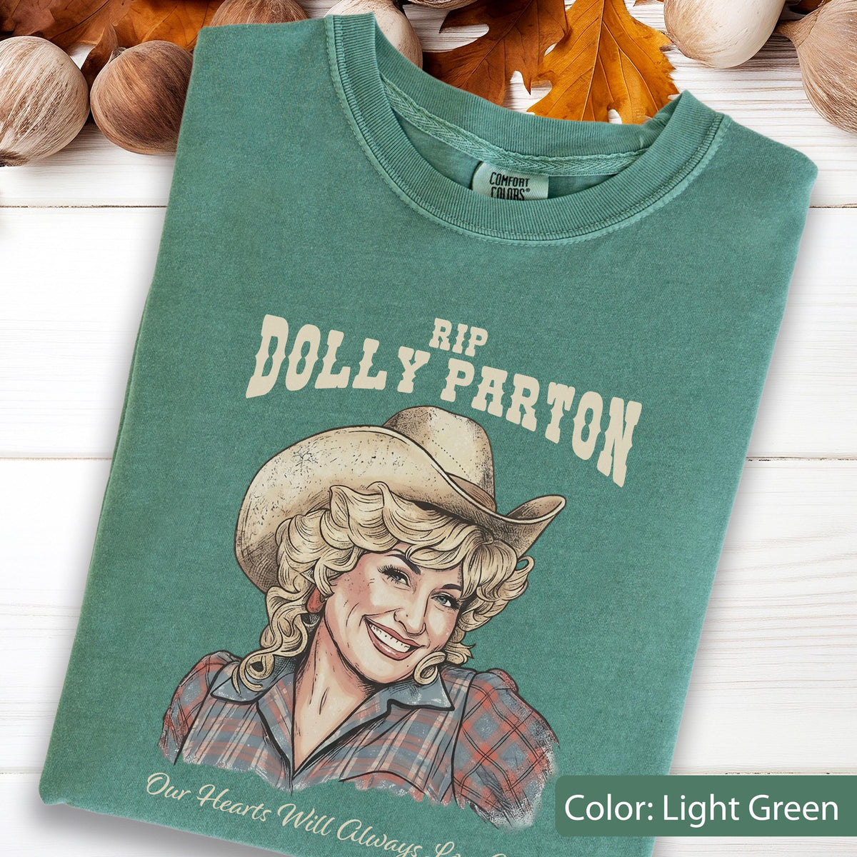 May include: A light green t-shirt featuring a graphic illustration of Dolly Parton wearing a cowboy hat. The text above reads RIP DOLLY PARTON and the text below reads Our Hearts Will Always Love You. The shirt has a crew neckline and a Comfort Colors label.