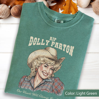 May include: A light green t-shirt featuring a graphic illustration of Dolly Parton wearing a cowboy hat. The text above reads RIP DOLLY PARTON and the text below reads Our Hearts Will Always Love You. The shirt has a crew neckline and a Comfort Colors label.