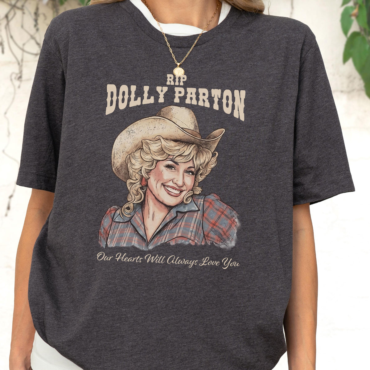May include: A dark heather gray short-sleeve t-shirt featuring a graphic illustration of Dolly Parton wearing a cowboy hat and a plaid shirt. The text reads RIP DOLLY PARTON at the top and Our Hearts Will Always Love You at the bottom.