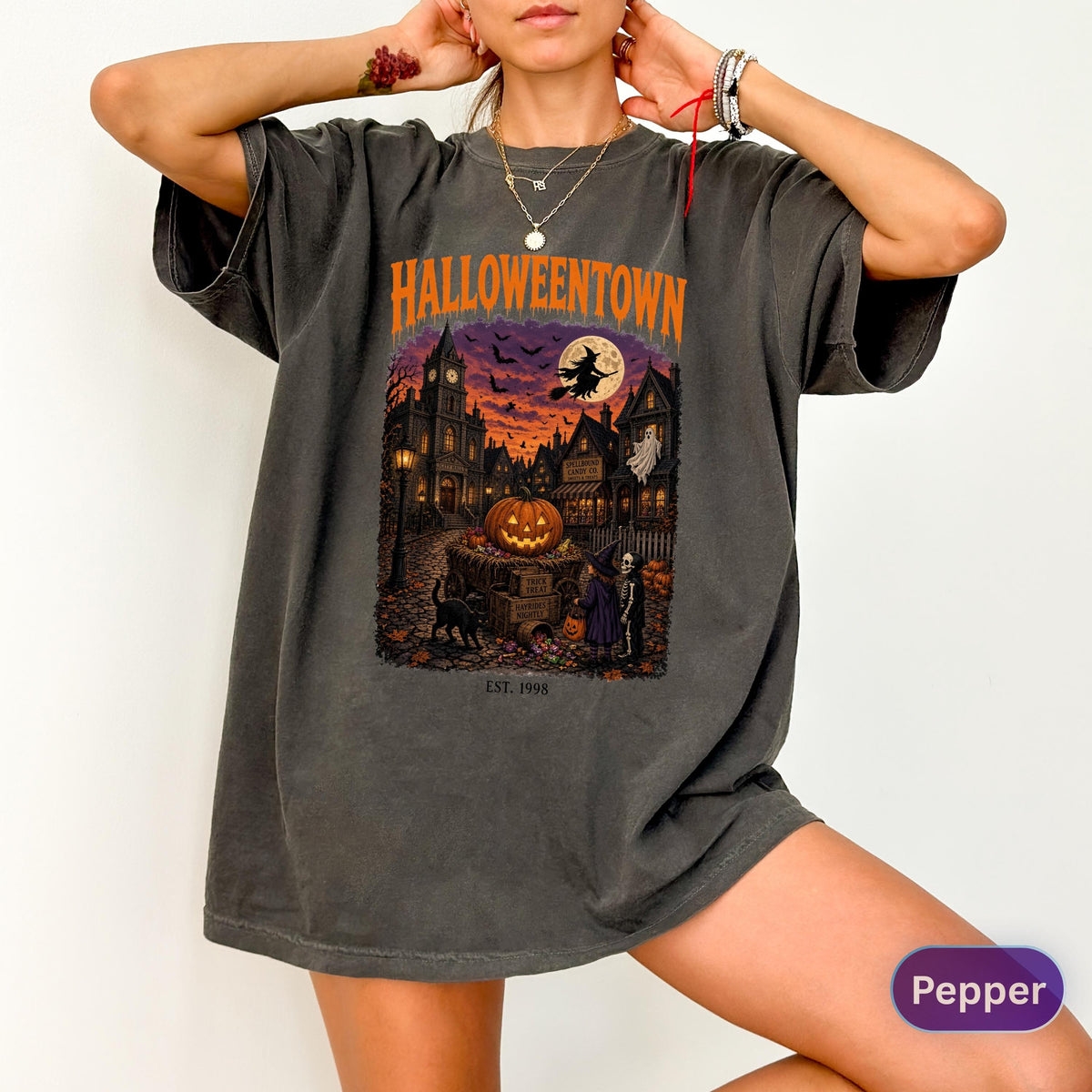 May include: A charcoal gray oversized t-shirt featuring a graphic print with the text HALLOWEENTOWN and EST. 1998. The illustration depicts a spooky town scene at night with a large pumpkin, a witch on a broomstick, a ghost, and a black cat.
