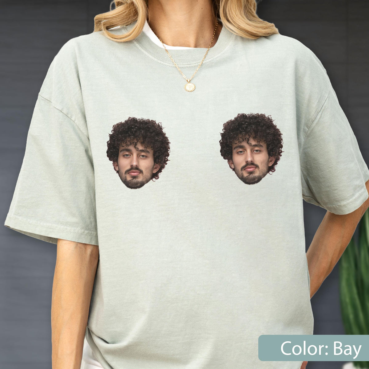 May include: A light sage green short-sleeve t-shirt featuring two identical photographic prints of a person with dark curly hair and a beard positioned side-by-side on the chest area. The shirt has a crew neckline and a relaxed fit.