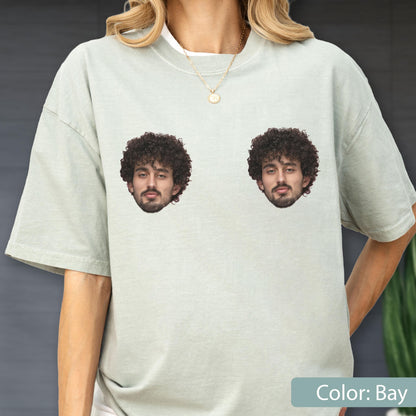May include: A light sage green short-sleeve t-shirt featuring two identical photographic prints of a person with dark curly hair and a beard positioned side-by-side on the chest area. The shirt has a crew neckline and a relaxed fit.