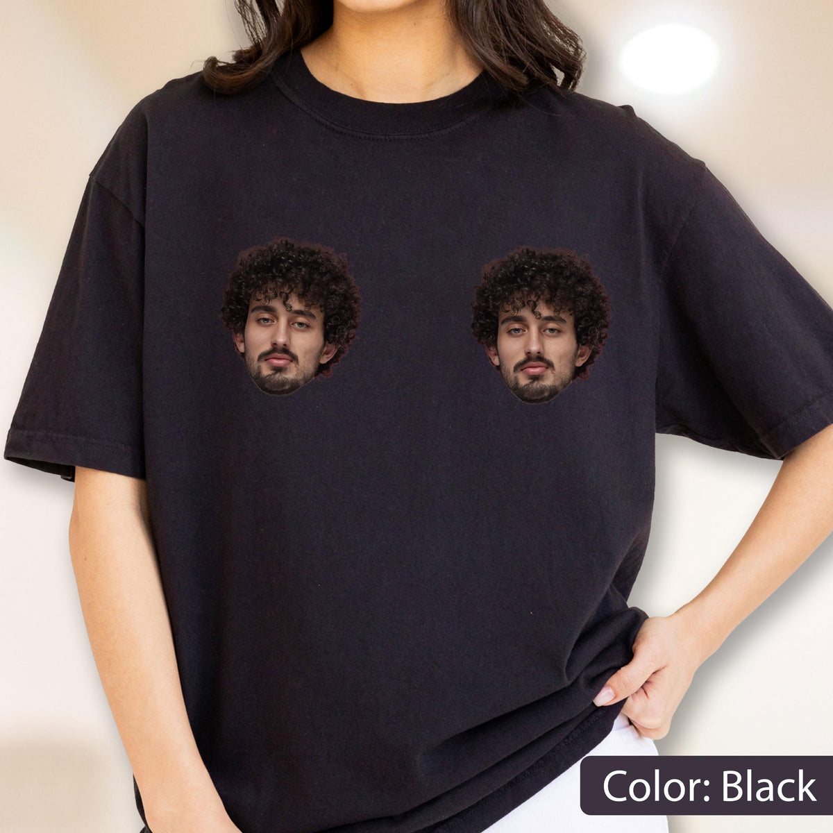 May include: A black short-sleeved t-shirt featuring two identical graphic prints of a person with dark curly hair and a beard positioned on the chest area. The shirt has a crew neckline and a standard fit, made from a solid black fabric.