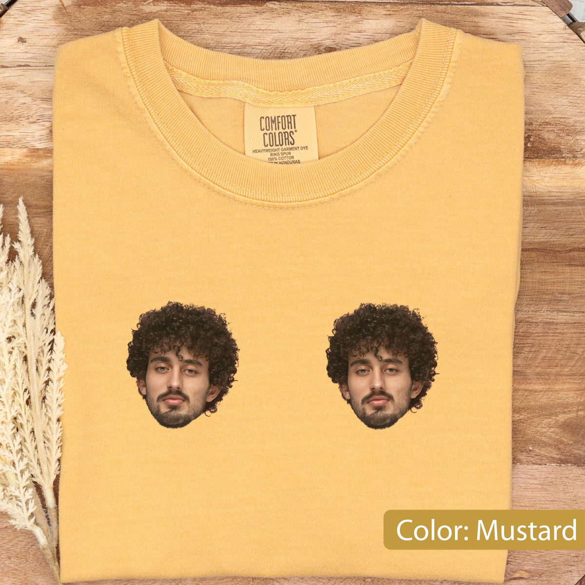 May include: A mustard yellow short-sleeve t-shirt featuring a crew neckline and a Comfort Colors brand label. The front of the shirt displays two identical graphic prints of a person with dark curly hair and a beard, positioned side by side.