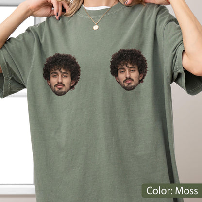 May include: A moss green crew neck t-shirt featuring two identical photographic prints of a person with dark curly hair and a beard. The graphic is positioned on the upper chest area of the garment, displaying the face from the chin up.