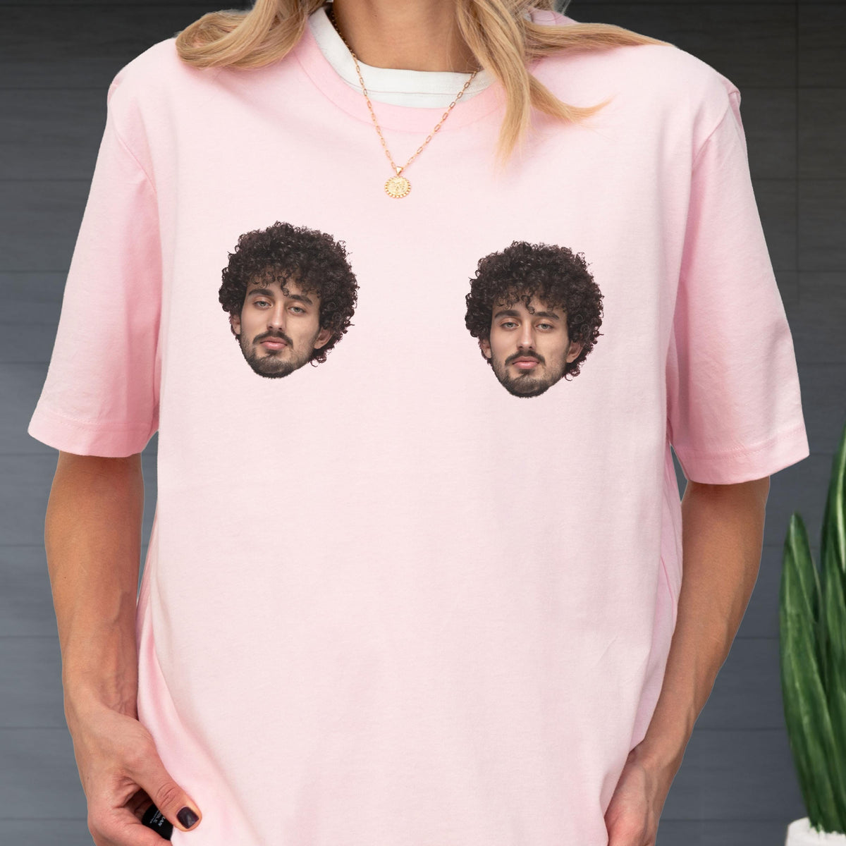 May include: A light pink short-sleeved t-shirt featuring two identical photographic prints of a person with dark curly hair and a beard positioned on the chest area. The shirt has a crew neckline and is worn with a thin gold chain necklace.