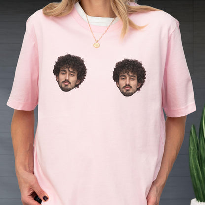 May include: A light pink short-sleeved t-shirt featuring two identical photographic prints of a person with dark curly hair and a beard positioned on the chest area. The shirt has a crew neckline and is worn with a thin gold chain necklace.