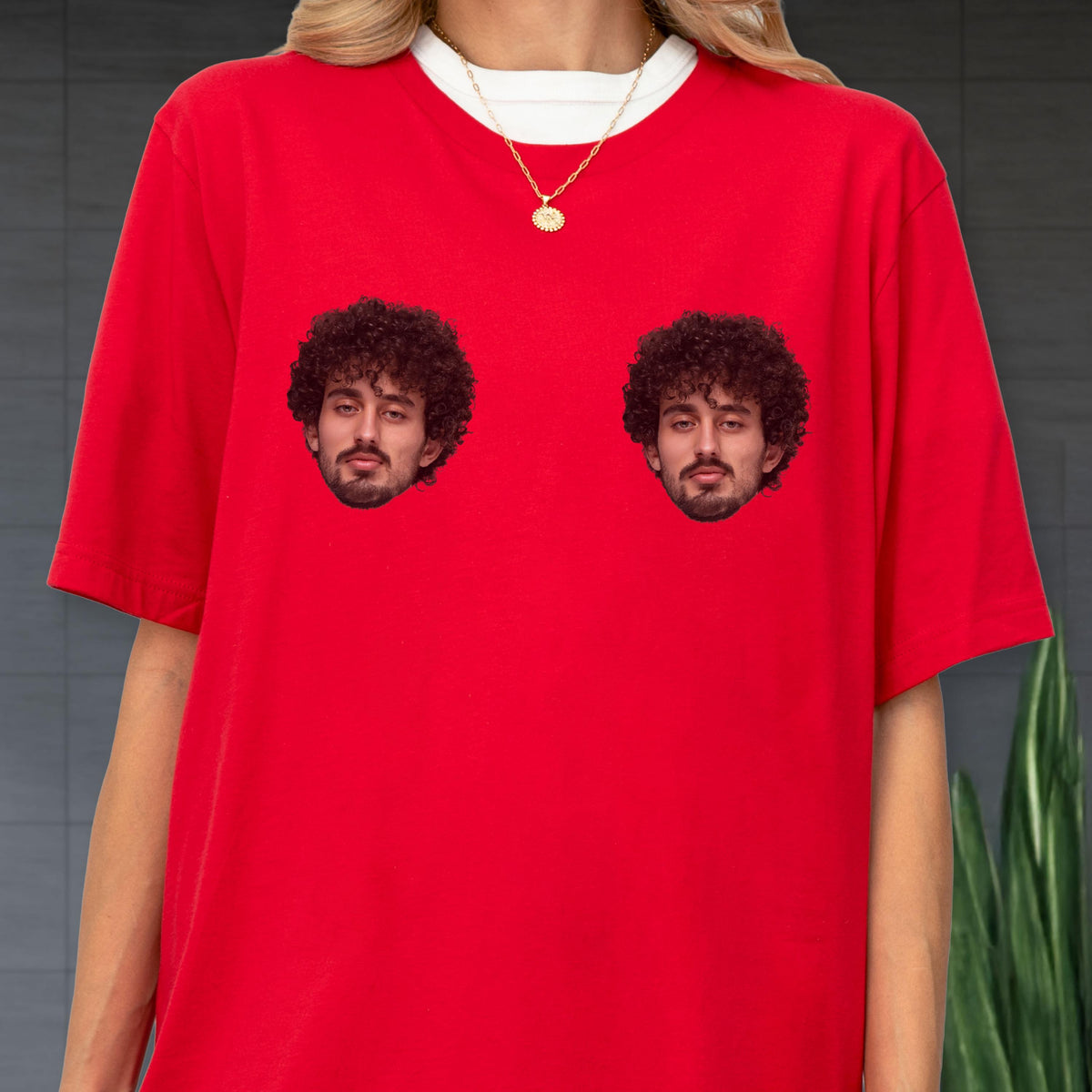 May include: A bright red short-sleeved t-shirt featuring two identical photographic prints of a person with dark curly hair and a beard positioned on the chest area. The shirt is worn over a white crew-neck undershirt with a gold chain necklace.