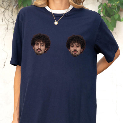 May include: A navy blue short-sleeved t-shirt featuring two identical photographic prints of a person with dark curly hair and a beard positioned on the chest area. The shirt has a crew neckline and is worn with a thin gold-toned chain necklace.
