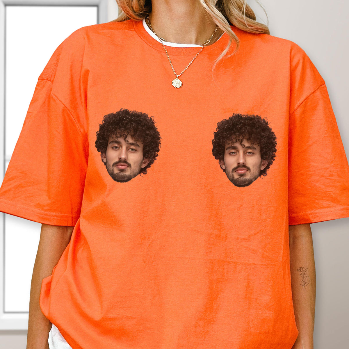 May include: A bright orange short-sleeved t-shirt featuring two identical graphic prints of a man's face with dark curly hair and a beard. The shirt has a crew neckline and a loose, relaxed fit, showcasing a vibrant solid color throughout the fabric.