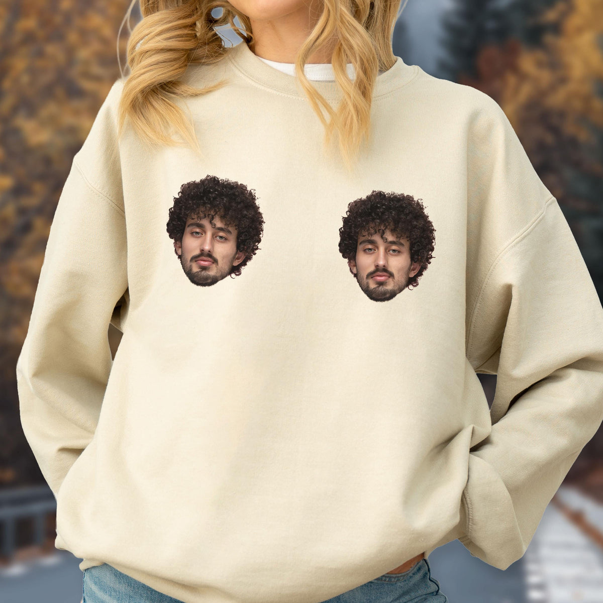 May include: A beige crewneck sweatshirt featuring two identical photographic prints of a person with dark curly hair and a beard positioned on the upper chest area. The garment has long sleeves, a ribbed neckline, and a relaxed fit throughout.