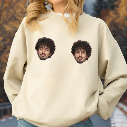 May include: A beige crewneck sweatshirt featuring two identical photographic prints of a person with dark curly hair and a beard positioned on the upper chest area. The garment has long sleeves, a ribbed neckline, and a relaxed fit throughout.