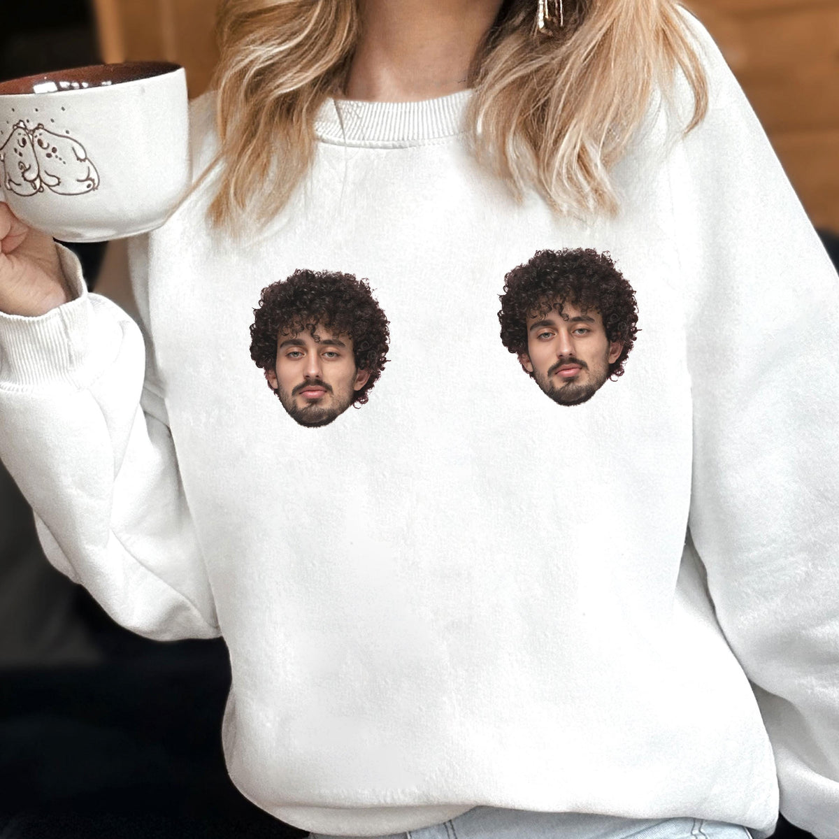 May include: A white crewneck sweatshirt featuring two identical photographic prints of a person with dark curly hair and a beard on the chest area. The sweatshirt is paired with a white ceramic mug decorated with a small line drawing of two bears.