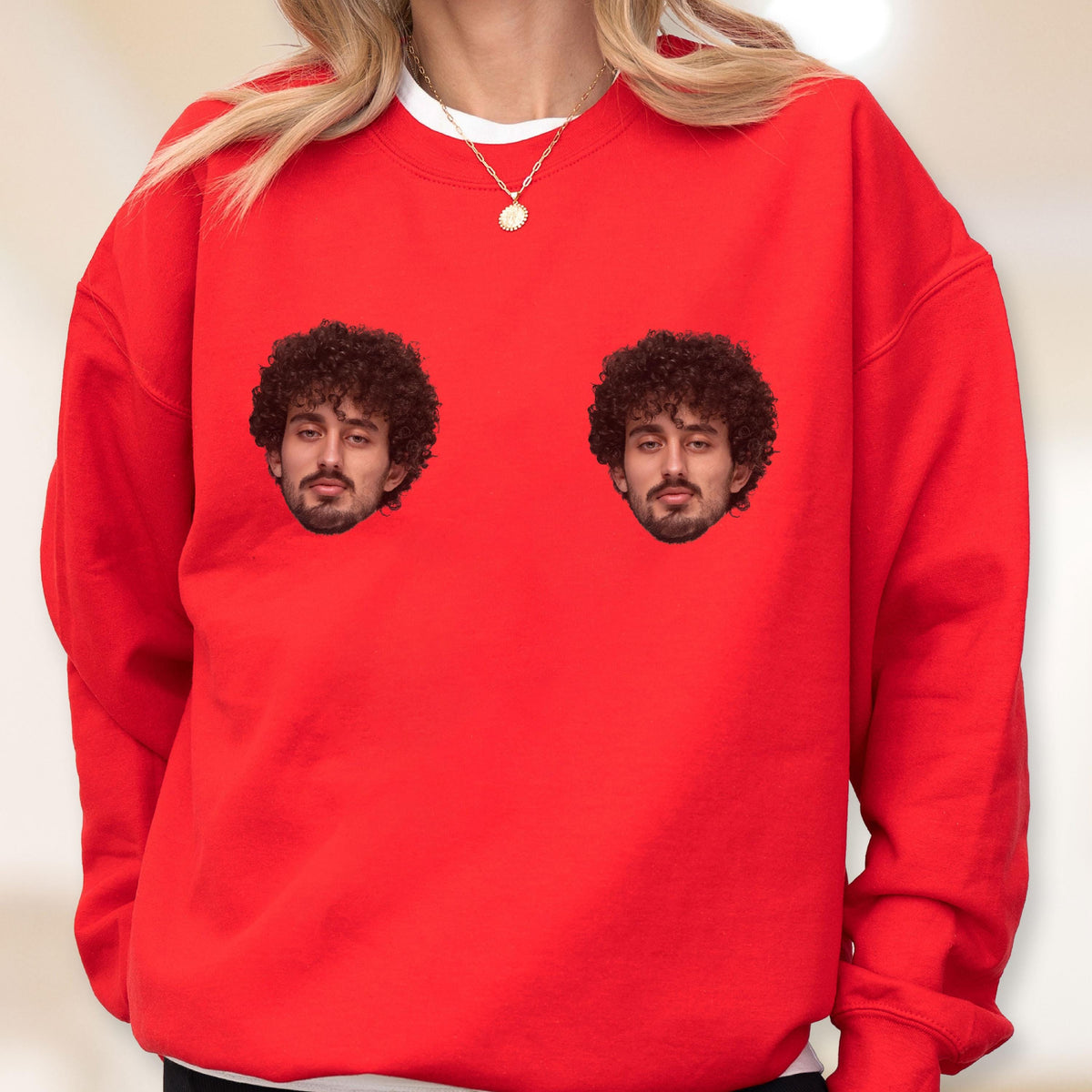 May include: A bright red crewneck sweatshirt featuring two identical graphic prints of a person with dark curly hair and a beard positioned on the chest area. The garment has long sleeves and a ribbed neckline, worn over a white undershirt.