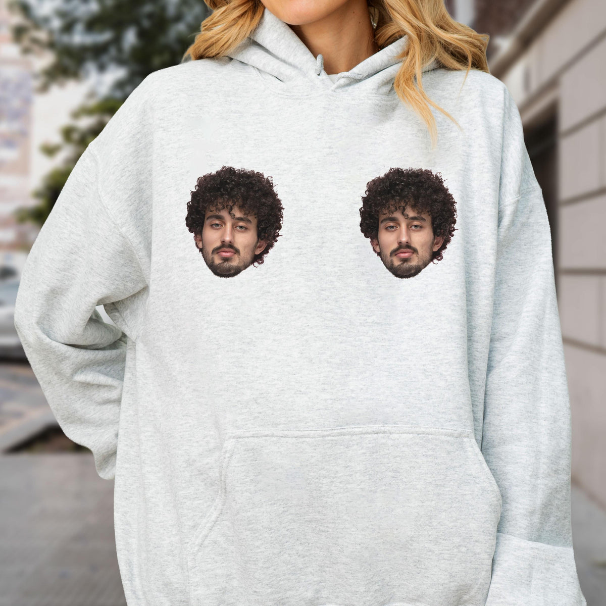 May include: A light gray heathered hooded sweatshirt featuring two identical graphic prints of a person with dark curly hair and a beard positioned on the chest area. The garment includes a hood with drawstrings and a large front kangaroo pocket.