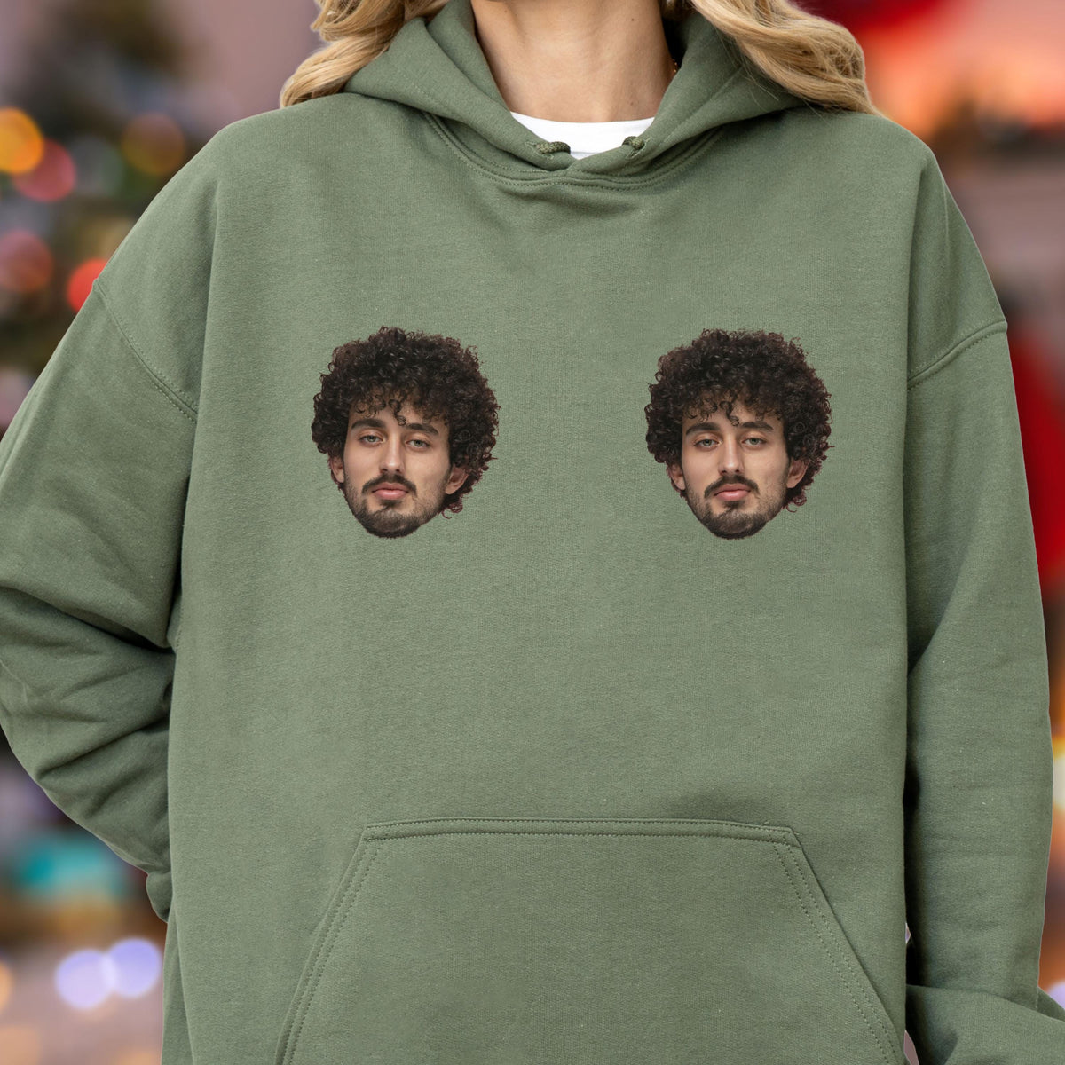 May include: An olive green hooded sweatshirt featuring two identical photographic prints of a person with dark curly hair and a beard positioned on the chest area. The garment includes a front kangaroo pocket and a drawstring hood.