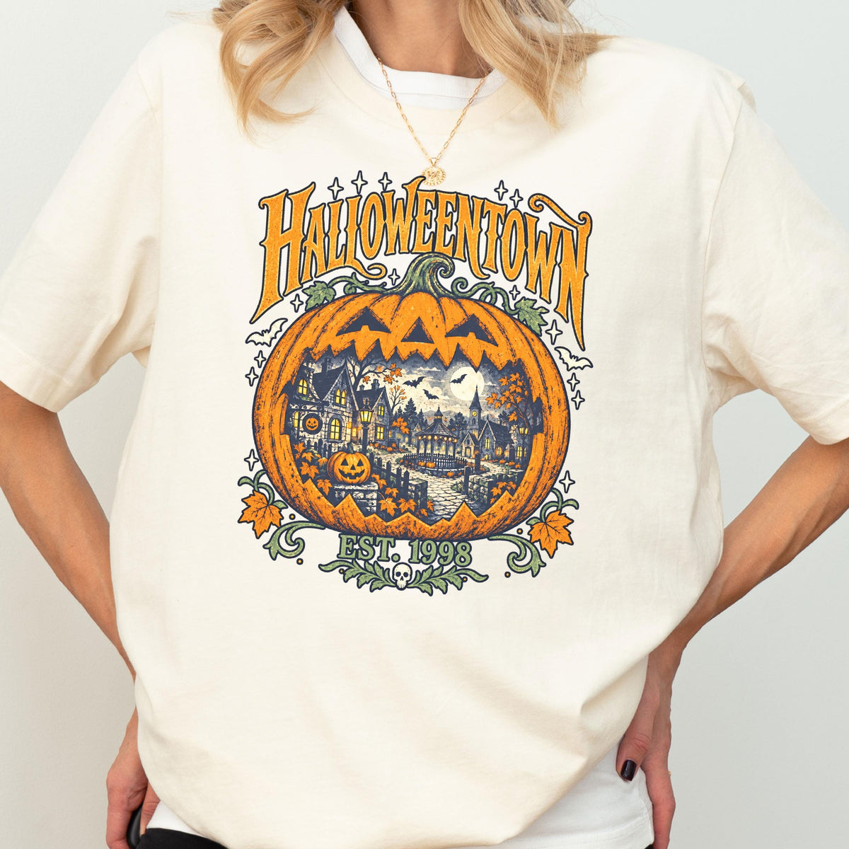 May include: A cream-colored t-shirt featuring a large orange jack-o'-lantern graphic. Inside the pumpkin is a detailed scene of a town with houses, trees, and bats. The text reads HALLOWEENTOWN and EST. 1998, with small stars and leaves surrounding it.